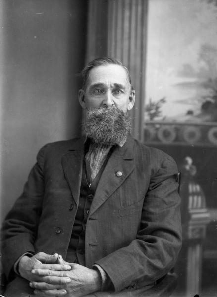 Studio portrait of a seated man with a curly moustache and beard in front of a painted backdrop. He has a pin on the lapel of his suit jacket, and is wearing a vest and necktie.