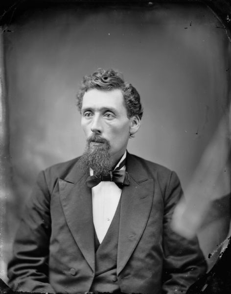 Studio portrait of a seated man in front of a painted backdrop with curly hair and a wiry moustache and beard, wearing a suit jacket, vest, and bow tie. Possibly Mr. Sly.	