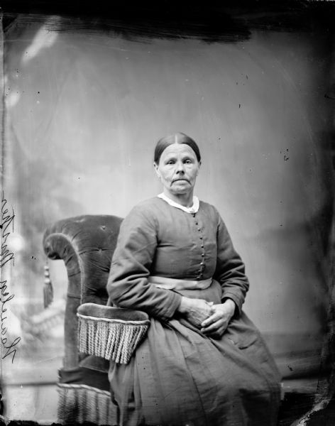 Studio portrait of a seated woman in a stuffed chair in front of a painted backdrop. Etching on negative says, "Heaselen's Mother."