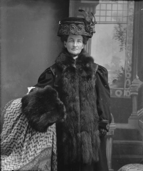Studio portrait of a standing woman wearing a fur coat and fur muff, possibly Julia Schnurr or Snurr.  Standing with draped support on left, with painted backdrop depicting stairway on right.  

