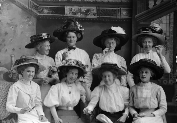 Studio portrait of eight women, four seated and four standing, all wearing hats and posing with cigarettes and cigars, in front of a painted backdrop. Top row, from left to right: Ella Perry, ? Ericksen, Maria Smick, and Mina Brandon. Bottom row, from left to right: Minnie Thompson, Sadie Lind, Fern Perry, and Martha Nelson.