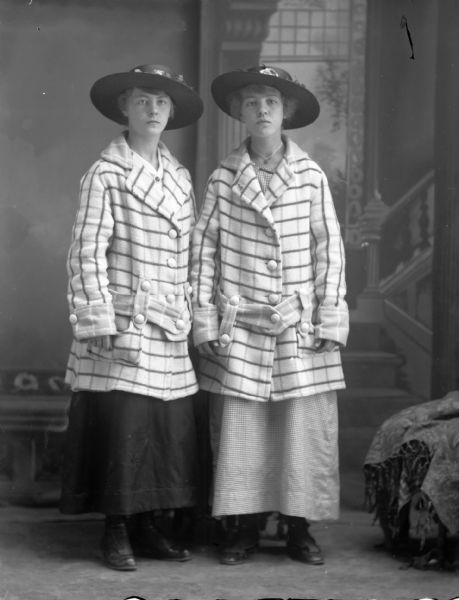 Studio portrait of two standing women wearing the same checked coat and hat, possibly the Gilbertsons, in front of a painted backdrop.	
