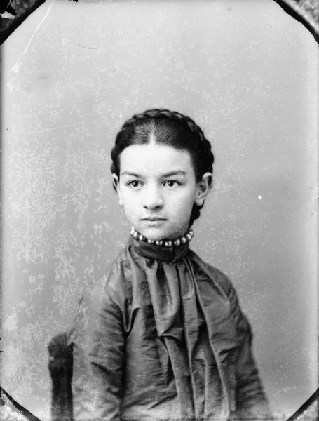 Studio portrait of a seated young woman with a braid circling her head.	She is wearing a necklace over a high-collared blouse.
