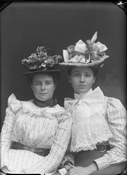 Studio portrait of two seated women wearing large hats. They are both wearing belts over their dresses, and the woman on the right has a large lace collar.