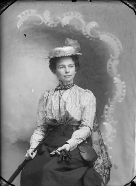 Studio portrait of a seated woman in front of a painted backdrop. She is wearing a hat and holding an umbrella in her right hand, possibly Effie Memholtz.