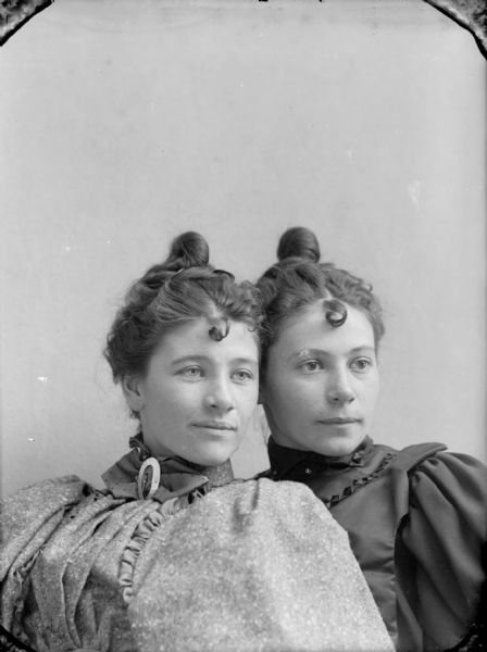 Studio portrait of two seated young women with matching hairstyles, including a single curl in the middle of their foreheads. They are both wearing puffy-sleeved blouses.