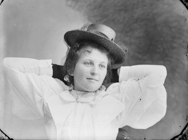 Studio portrait of a seated young woman wearing a hat with a veil, gloves, and a puffy-sleeved blouse, posing with her hands behind her head.