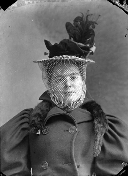Studio portrait of a seated young woman wearing a hat with a veil and flower, a mink skin placed around the collar, and a double-breasted coat with gathered, tucked sleeves. She is facing forward, with her head tilted to the right.