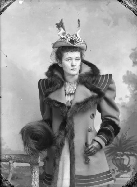 Studio portrait of a standing young woman, wearing a hat with a veil and flower, and a fur-lined coat in front of a painted backdrop.