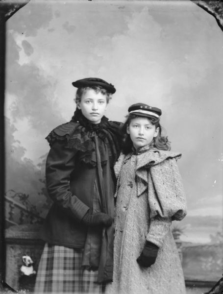 Studio portrait of two girls wearing winter coats, gloves, and hats in front of a painted backdrop.
