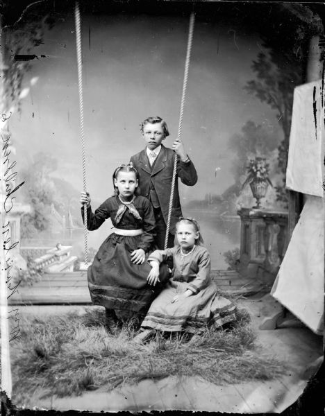 Studio portrait of two girls and a boy posed with a swing in front of a painted backdrop. One girl sitting on the swing, one girl sitting on the ground, and the boy standing. Etching on negative says "Wm. Hearlen Children - 6."