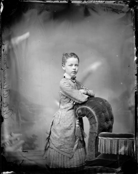 Studio portrait of a girl standing and leaning on the back of a stuffed chair in front of a painted backdrop. Probably Joan Olson, the daughter of Pete Olson.