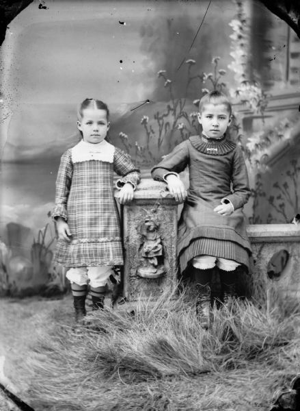 Studio portrait of two small girls leaning on a stone fence, with one sitting on the fence in front of a painted backdrop.