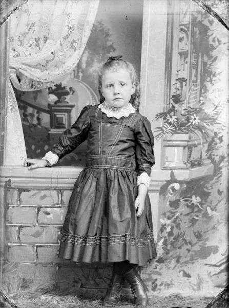 Studio portrait of a girl in a dress standing with her legs crossed, leaning on a brick wall in front of a painted backdrop.