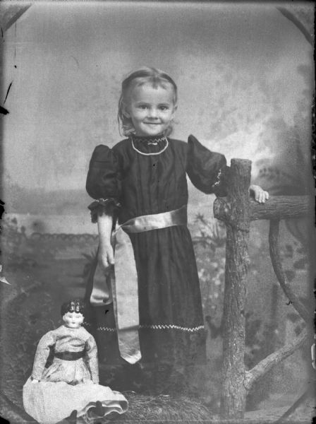 Studio portrait of a girl, in a dress with a sash around the waist, standing with her hand on a wooden fence near a doll sitting on the ground in front of a painted backdrop.