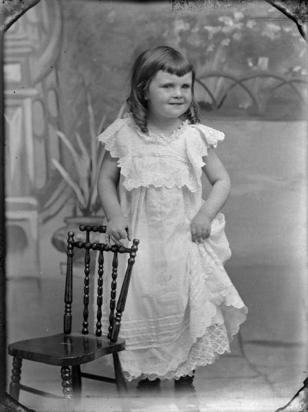 Studio portrait of a girl standing in a white dress, holding up a part of her dress and resting her hand on a chair in front of a painted backdrop.	
