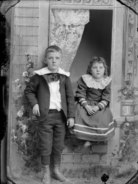Studio portrait of a girl sitting on a stone window ledge and a boy standing. The window and wall are a painted prop.