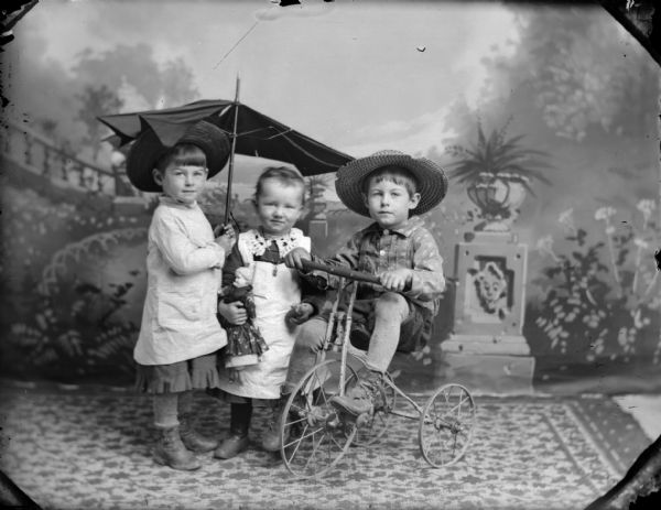 Studio portrait of three children posing in front of a painted backdrop. Two children are standing, and one holds an umbrella and the other a doll. The other child sits on a tricycle.