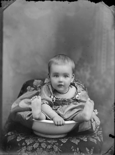 Studio portrait of a baby with bare feet sitting in a bowl in front of a painted backdrop.	
