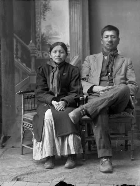 Full-length studio portrait of Thomas Thunder (Hoonk Ha Ga Kah) (Son of [Wa Con Cha Kah] John Thunder aka Dr. Thunder and [We Hon Pe Kaw] Lucy Bear, Thunder), and his wife, Addie Littlesoldier, Lewis, Thunder (Kzunch Je Kay Ra Win Kah) (Daughter of [No Gin Kaw] Little Soldier and Mrs. Cho Ne We Kaw). He is wearing a scarf around his neck, with a suit jacket, vest, and trousers. She is wearing a coat over a dress. In the background is a painted backdrop.