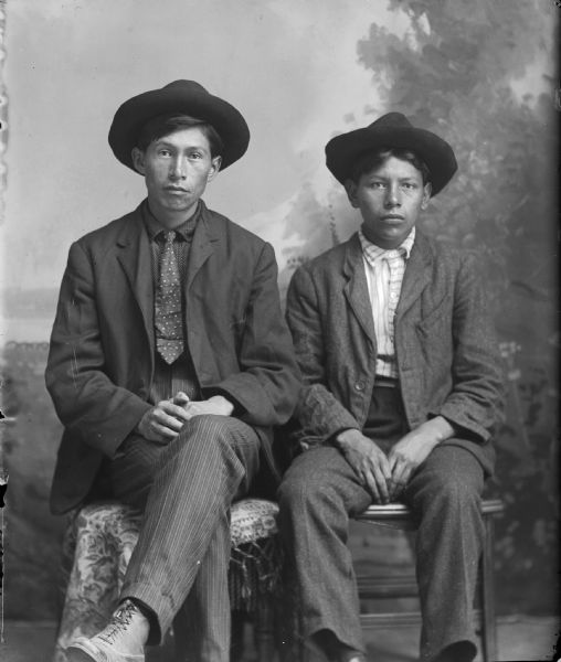 Studio portrait of two seated young Native American men in hats in front of a painted backdrop. They are both wearing suit jackets, vests, and trousers. The man on the left is wearing a necktie.