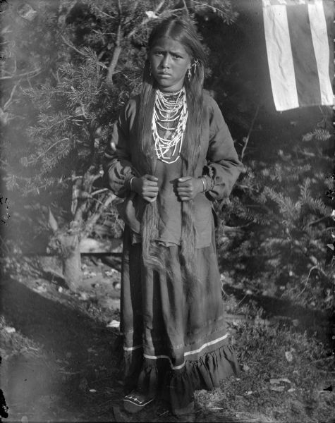 Belle Mattie Mike "ENooKah" (First Daughter), daughter of John Mike, Jr. "HayShooKeeKah" (One Horn) and his wife, Kate Littleblackhawk "ENooNeeKah" (Woman). She is posing outdoors, in front of pine trees, wearing indigenous apparel.