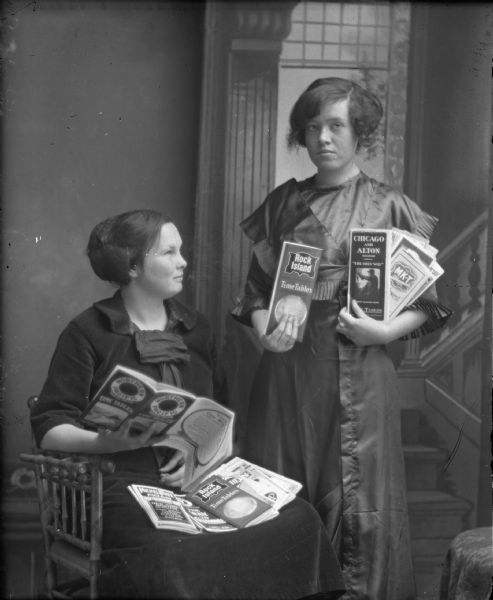 Studio portrait of two women, one standing and the other seated, displaying train time tables in front of a painted backdrop.