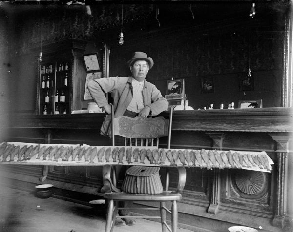 Moses Pauquette shows off his catch of fish with his fishing creel at his tavern on Water Street. He stands before the bar, leaning on his left arm. His catch of 44 fish are lined up on a tray, balanced on the arms of a chair and his creel, in front of him.