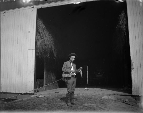 Fisherman with a pole and a fish on a line in front of an open barn doorway.