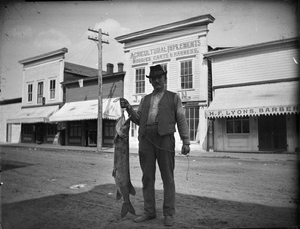Man, probably Ira Bowen, holding a muskellunge in front of H.F. Lyon's barbershop on Water Street, between Main and Harrison Streets.	