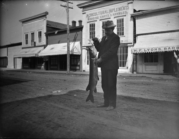Man, probably Ira Bowen, holding a muskellunge in front of H.F. Lyon's barbershop on Water Street, between Main and Harrison Streets.