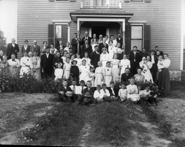 Large group of men, women, and children, probably a wedding, standing in front of a house.
