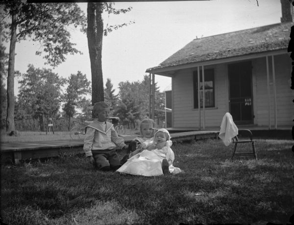 Boy and girl sitting on the grass next to the board sidewalk in front of a house. The girl is also holding an infant and dog, possibly Dorothy Jones (later Krohn), and Douglas York, a cousin, and the infant W.P. Jones.