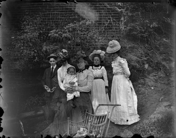 Four women, man, and infant posed standing in front of a brick wall.