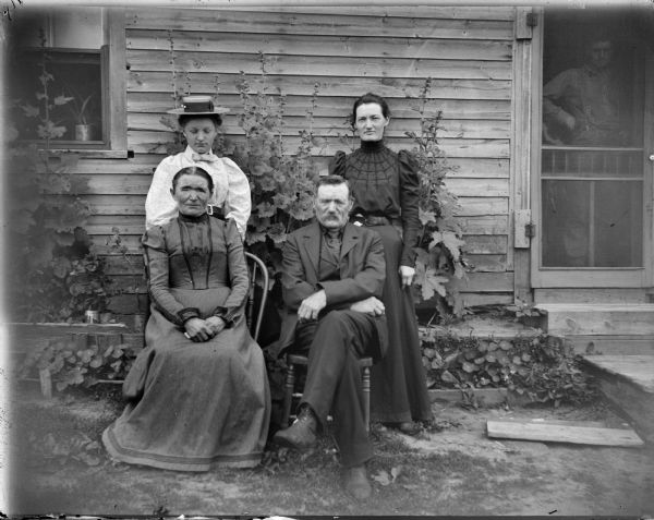 Two women stand behind an elderly man and woman seated in front of a frame house. There is a man visible through the screen door.