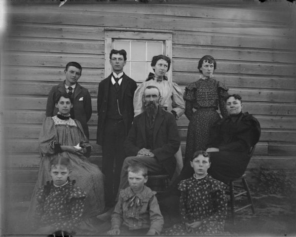 Two men and a women seated in chairs, two young men and two young women standing behind the chairs, and two girls and a boy seated in front of the chairs, all posing in front of a house.