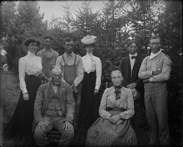 Elderly woman and man seated, surrounded by four men and two women, probably the Phillip Nortman family, posing among fir trees. Reportedly, both Nortman girls went to Milwaukee, circa 1907-1908, and married/divorced African American men.