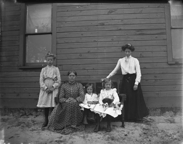 A woman and two girls are sitting on a wooden bench in front of a house, and another woman and girl are standing alongside the bench. From left to right: possibly Aleda Johnson, Sarah Johnson Halverson, Lenore Johnson, Beatrice Johnson, and Alma Halverson Horswill. Alma Horswill was the daughter of Sara Halverson, and the girls were nieces of the latter.