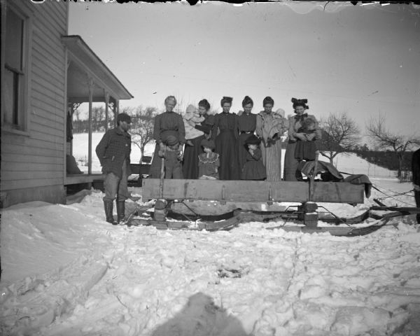 Winter scene with six women, six children, and a man examining a bobsled hitched to a horse.