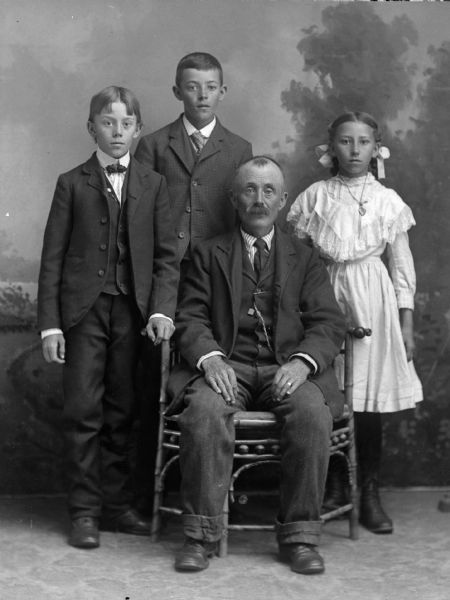 Studio portrait of a seated man surrounded by two standing boys and a girl in a dress in front of a painted backdrop. The man and boys are wearing suit jackets, neckties, vests, and trousers.