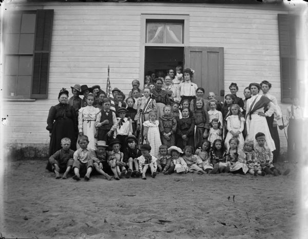 Group portrait of a large group of people, primarily children, gathered around the doorway and outside of a building with a United States flag.