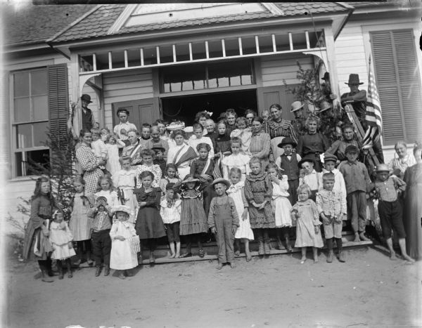 Group portrait of a large group of people, primarily children, gathered on the porch of a building with a United States flag.