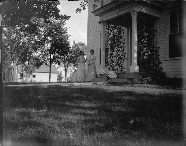 Two women standing under a small parasol/umbrella in front of a house numbered 1102, probably the home of W.T. Murray at the corner of Tenth and Main Streets, and a small girl seated on the steps of the house.	

