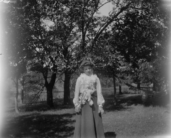 Woman posed standing in a yard among deciduous trees.	