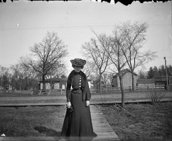 Woman wearing a fur stole, and posed standing on a board sidewalk.