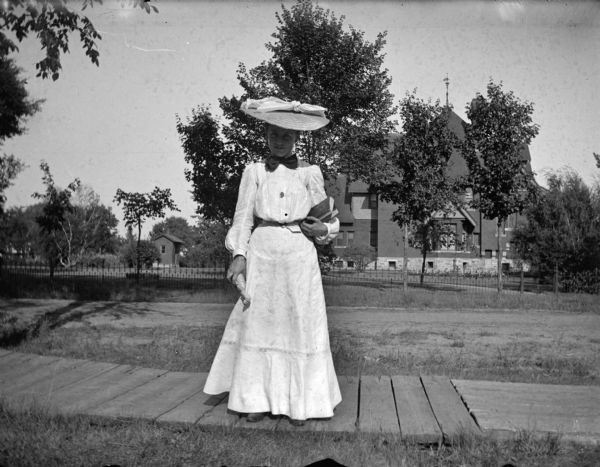 Woman with books posed standing on a board sidewalk, possibly in front of the William T. Price home.
