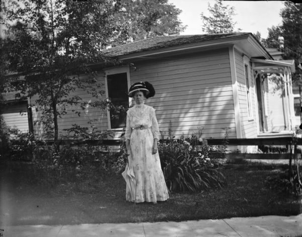 Woman posed standing with a folded umbrella by a flower bed in the yard of a house.