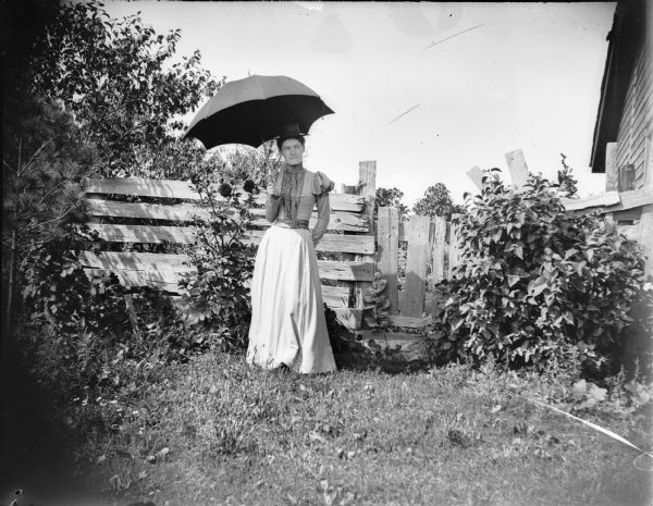 Woman posed standing with an open umbrella in a flower garden by a wooden fence and gate.	