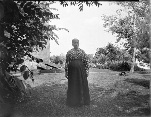 Elderly woman in a striped blouse posed standing in a yard.	