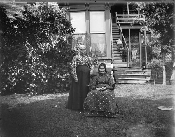 Two elderly women posed standing and sitting in a yard.